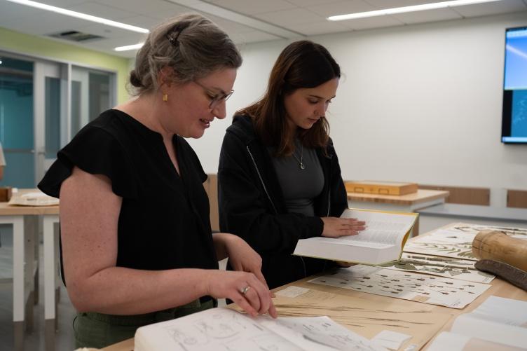 A photo showing two women standing at a table and looking through specimens from the Hodgdon Herbarium of an extinct crabgrass found only in New Hampshire at one time.